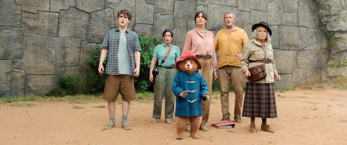 Paddington und seine Familie stehen vor einer Steinwand und gucken mit großen Augen etwas entgegen. 