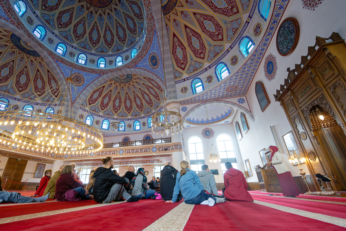 Am Tag der offenen Moschee sitzen Besucherinnen und Besucher bei einer Führung in der Merkez Moschee in Duisburg auf dem Teppich im Gebetsraum. 